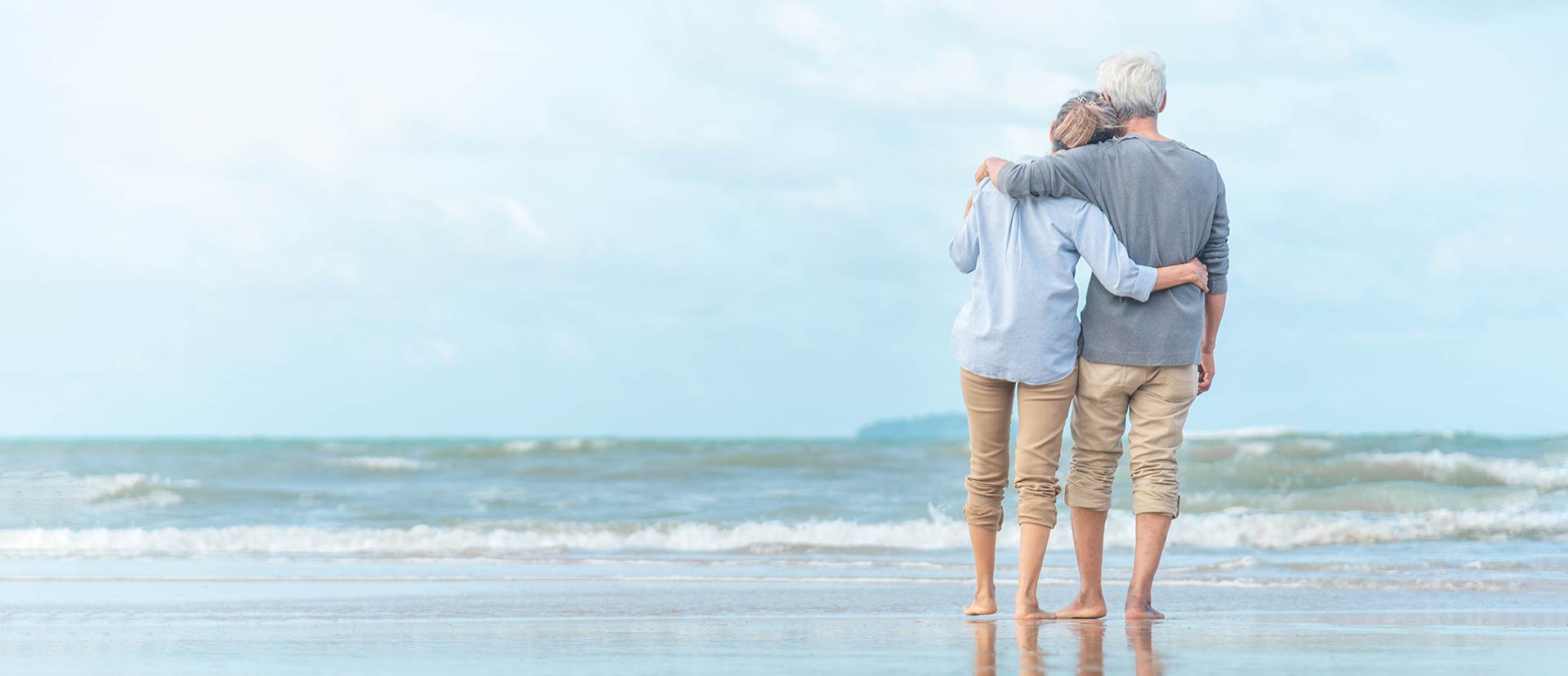 Ein älteres Paar steht Arm-in-Arm am Strand und schaut aufs Meer