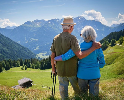Ein älteres Ehepaar steht vertraut umschlungen auf einer sonnigen Bergwiese und genießt gemeinsam den weiten Blick über eine atemberaubende Alpenlandschaft. Der Mann mit beigem Hut und olivgrünem Hemd hat seinen Arm liebevoll um die Frau in blauer Jacke gelegt, während er sich entspannt auf seinen Wanderstock stützt. Vor ihnen erstreckt sich ein Panorama aus saftig grünen Almwiesen, dichten Nadelwäldern und majestätischen schneebedeckten Gipfeln unter einem leuchtend blauen Himmel mit weißen Wolken. Das Bild strahlt eine tiefe Ruhe und Gelassenheit aus - es zeigt, wie das Paar unbeschwert die Natur und ihre gemeinsame Zeit auf Reisen genießt, ohne sich Sorgen machen zu müssen.