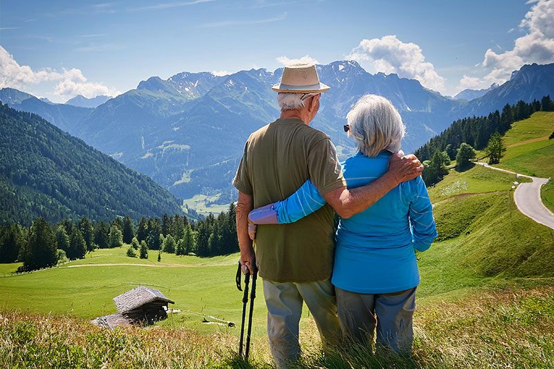 Ein älteres Ehepaar steht vertraut umschlungen auf einer sonnigen Bergwiese und genießt gemeinsam den weiten Blick über eine atemberaubende Alpenlandschaft. Der Mann mit beigem Hut und olivgrünem Hemd hat seinen Arm liebevoll um die Frau in blauer Jacke gelegt, während er sich entspannt auf seinen Wanderstock stützt. Vor ihnen erstreckt sich ein Panorama aus saftig grünen Almwiesen, dichten Nadelwäldern und majestätischen schneebedeckten Gipfeln unter einem leuchtend blauen Himmel mit weißen Wolken. Das Bild strahlt eine tiefe Ruhe und Gelassenheit aus - es zeigt, wie das Paar unbeschwert die Natur und ihre gemeinsame Zeit auf Reisen genießt, ohne sich Sorgen machen zu müssen.
