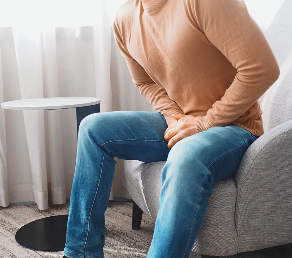 Ein Mann in Jeans und beigem Pullover sitzt auf einem Ohrensessel und presst die Hände in den Schritt, als hätte er Schmerzen oder muss dringend auf die Toilette.