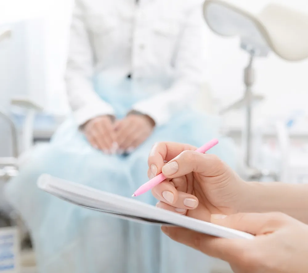 Im Vordergrund sieht man die Hände einer Ärztin, die auf einem Zettel eine Diagnose notiert. Im Hintergrund erkennt man unscharf einen Urologie-Stuhl und einen darauf sitzenden Patienten. Die Atmosphäre ist hell und angenehm.