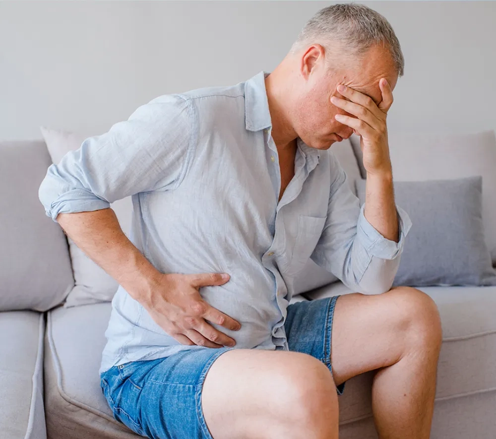 Ein Mann mittleren Alters sitzt auf einer hellen Couch und hält sich den Bauch vor Schmerzen. Den Kopf hat er verzweifelt in die Hand gestützt.