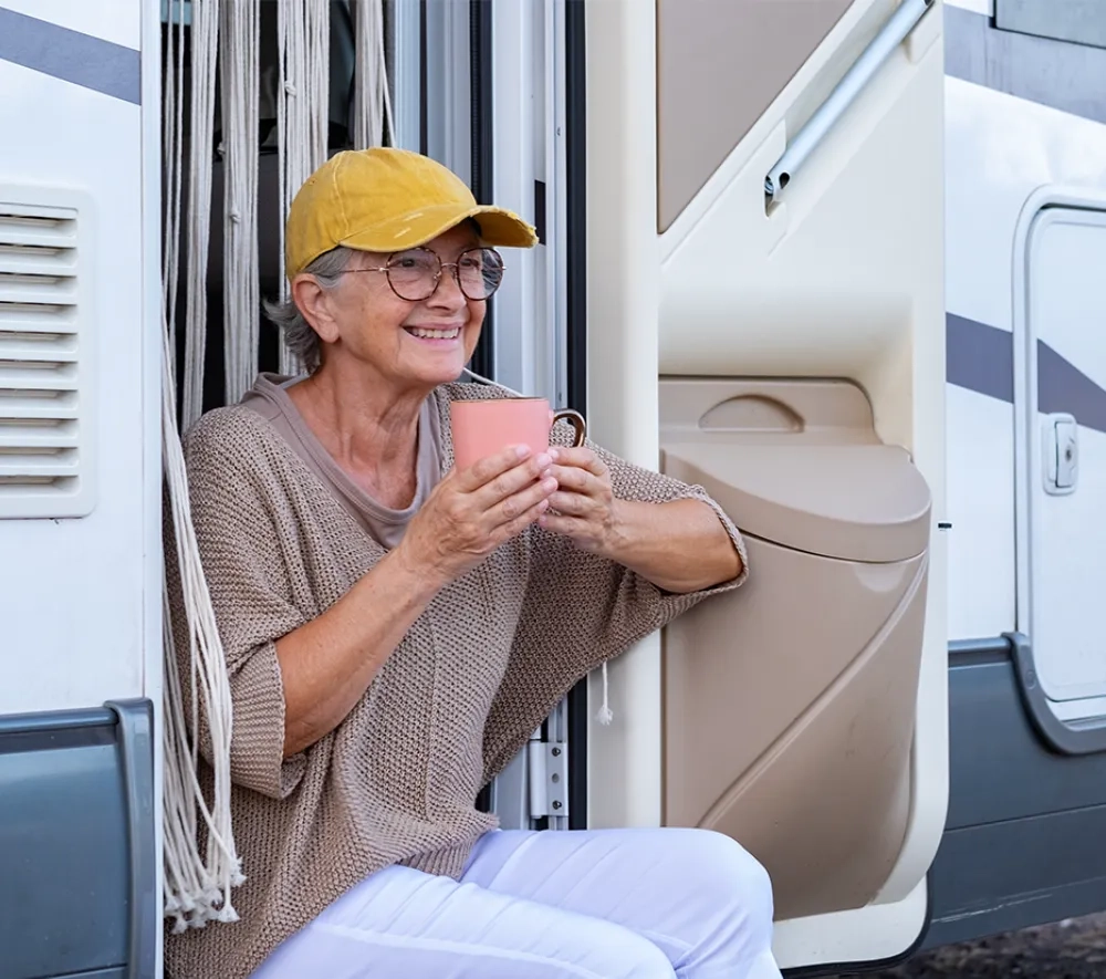 Eine ältere Dame mit grauem Haar und gelber Baseballkappe sitzt entspannt in einem modernen Wohnmobil und genießt sichtlich ihren Kaffee aus einer rosa Tasse. Sie trägt eine beige Strickjacke und eine helle Hose und lächelt warmherzig in die Kamera. Hinter ihr ist die saubere, helle Innenausstattung des Wohnmobils zu sehen mit weißen Schränken und Jalousien. Die Szene strahlt pure Lebensfreude und Unbeschwertheit aus - hier ist eine Frau, die das Reisen und ihre Freiheit in vollen Zügen genießt. Das Bild vermittelt das Gefühl von Sicherheit, Komfort und der Gewissheit, dass auch im Alter Abenteuer und spontane Reisen möglich sind, ohne sich Sorgen machen zu müssen.