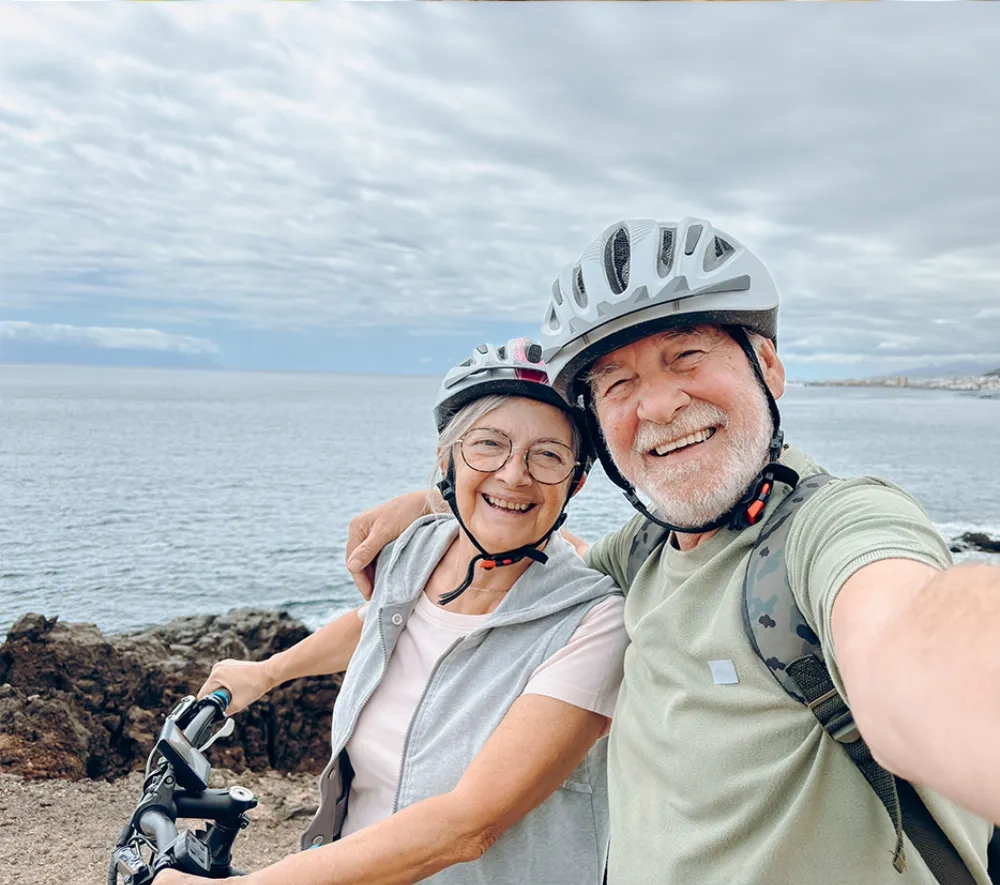 Ein älteres Ehepaar macht ein fröhliches Selfie während einer gemeinsamen Fahrradtour am Meer. Beide tragen weiße Fahrradhelme und strahlen vor Lebensfreude - die Frau mit grauer Kurzhaarfrisur und Brille lächelt herzlich, während der Mann mit grauem Vollbart ausgelassen lacht. Sie umarmen sich liebevoll, im Hintergrund erstreckt sich das weite, bläuliche Meer unter einem bewölkten Himmel. Das Bild vermittelt pure Unbeschwertheit und Lebensqualität - zwei Menschen, die trotz ihres Alters aktiv und voller Energie gemeinsame Abenteuer erleben, ohne sich durch körperliche Einschränkungen davon abhalten zu lassen. Die spontane, glückliche Stimmung zeigt: Mit den richtigen Hilfsmitteln ist ein aktives, sorgenfreies Leben in jedem Alter möglich.
