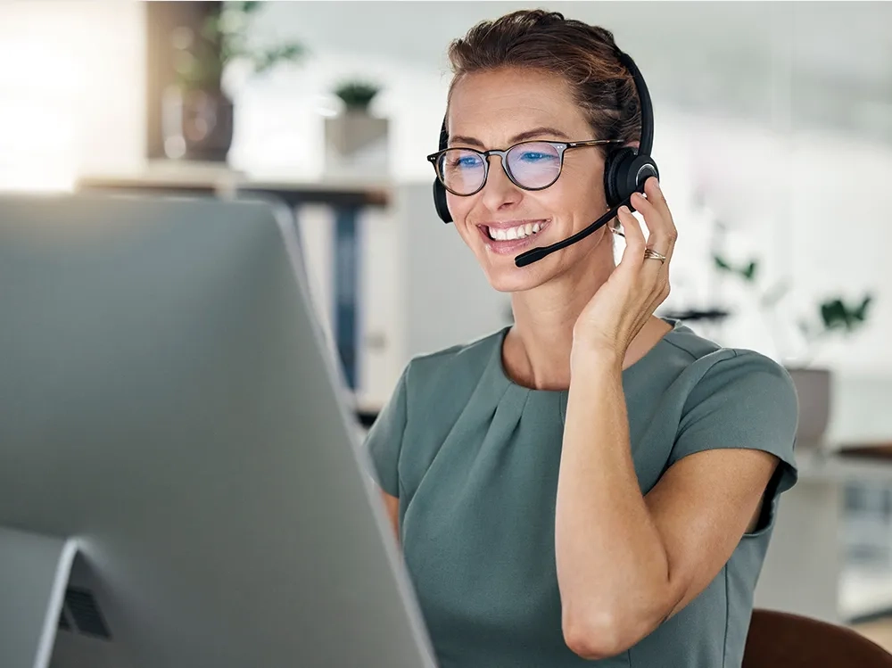 Eine freundlich lächelnde Servicemitarbeiterin sitzt vor Ihrem Computer und spricht in ihr Headset.