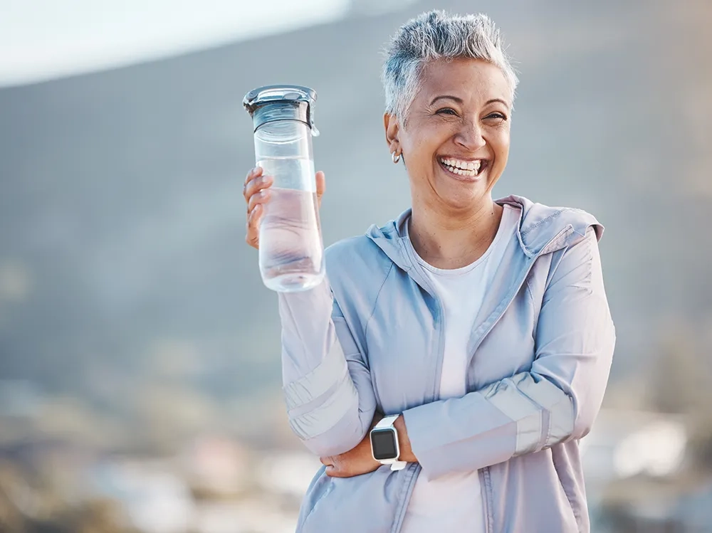 Eine vitale Frau mittleren Alters mit kurzem grauem Haar strahlt pure Lebensfreude aus, während sie nach dem Sport selbstbewusst eine Wasserflasche in die Höhe hält. Ihr herzliches, unbekümmertes Lachen und ihre entspannte Sportkleidung vermitteln das Gefühl grenzenloser Freiheit und Unbeschwertheit. Die warme Beleuchtung und der unscharfe Hintergrund unterstreichen ihre positive Ausstrahlung und Energie.