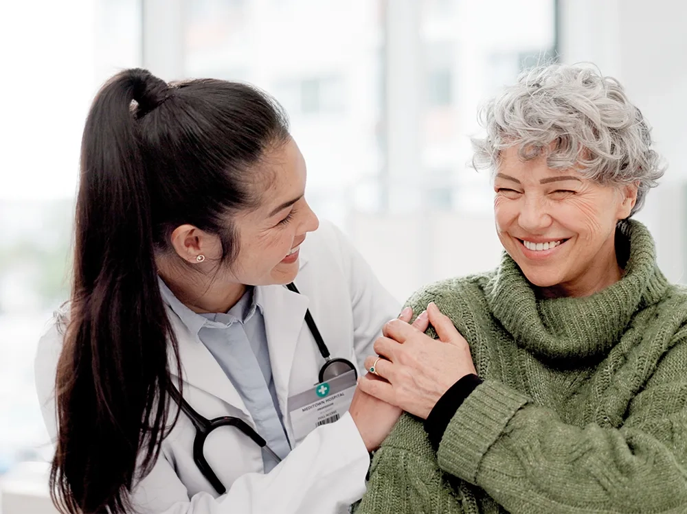 Eine junge Ärztin mit dunklen, zu einem Zopf gebundenen Haaren und weißem Kittel wendet sich warmherzig einer älteren Patientin zu. Die Patientin, eine Frau mit kurzen, silbergrauen Locken, trägt einen gemütlichen olivgrünen Strickpullover und strahlt voller Lebensfreude. Beide Frauen lächeln einander herzlich an - die Ärztin mit fürsorglicher Zuwendung, die Patientin mit einem strahlenden, selbstbewussten Lächeln, das pure Lebensfreude ausstrahlt. Die Patientin hat ihre Hand vertrauensvoll auf die Schulter der Ärztin gelegt, was die innige, vertrauensvolle Beziehung zwischen ihnen unterstreicht. Im Hintergrund ist eine helle, freundliche Praxisumgebung zu erkennen. Das Bild vermittelt Geborgenheit, Vertrauen und vor allem die Botschaft, dass ein erfülltes, sorgenfreies Leben trotz gesundheitlicher Herausforderungen möglich ist - die ältere Dame wirkt selbstsicher, aktiv und voller Optimismus.