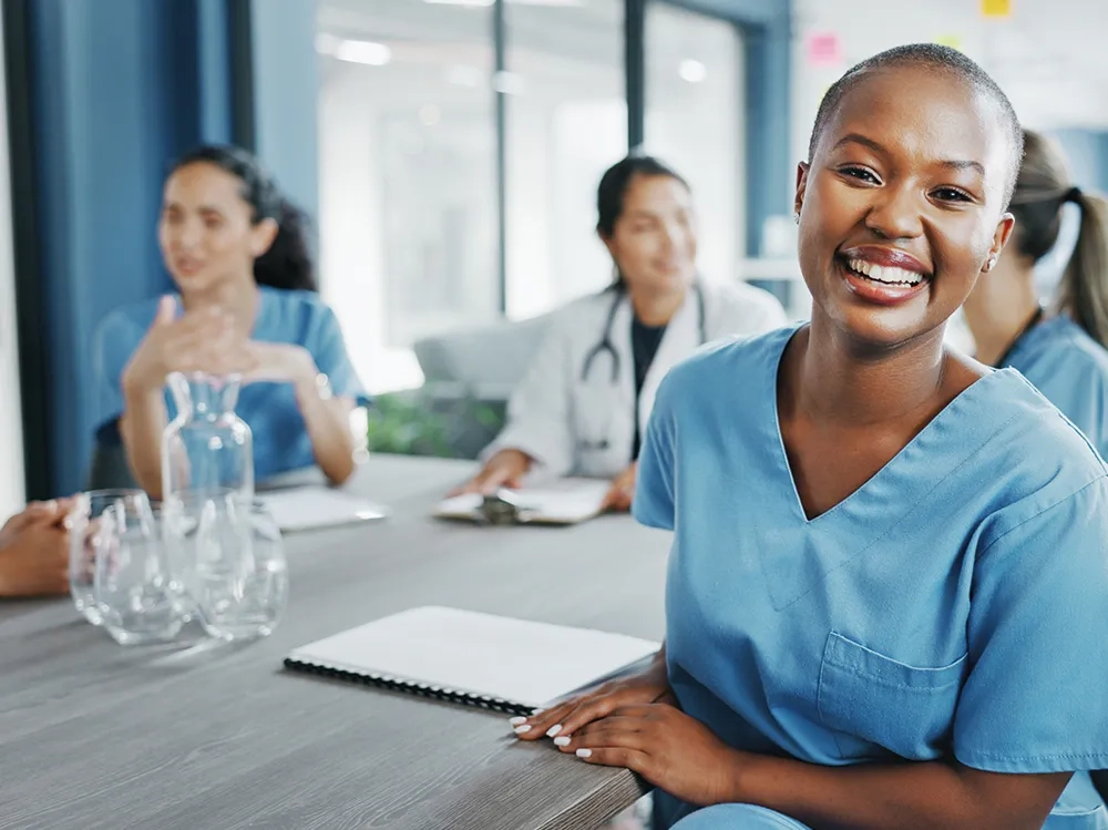 Eine junge, lächelnde Krankenschwester mit kurzen Haaren und hellblauer Arbeitskleidung steht strahlend im Vordergrund eines modernen Schulungsraums. Ihr offenes, herzliches Lachen und ihre entspannte Körperhaltung vermitteln Selbstvertrauen und Professionalität. Im Hintergrund sitzen weitere Kollegen in medizinischer Arbeitskleidung an einem Tisch - eine Frau in Blau und ein Arzt im weißen Kittel - die ebenfalls freundlich und engagiert wirken. Die helle, einladende Atmosphäre des Raums mit großen Fenstern unterstreicht das Gefühl von Kompetenz und Vertrauen. Das Bild strahlt aus: Hier arbeiten qualifizierte Fachkräfte, die durch fundierte Schulungen bestens darauf vorbereitet sind, Menschen mit Kontinenzproblemen einfühlsam und professionell zu betreuen - für ein Leben voller Würde und Unbeschwertheit.