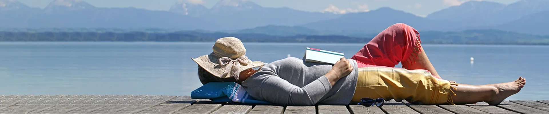 Eine Frau liegt auf dem Rücken auf einem sonnigen Holzsteg am Seeufer und genießen einen friedlichen Moment. Sie trägt einen grauen Pullover und eine bunte Hoste, hat ihren Sonnenhut über das Gesicht gelegt und ihr aufgeschlagenes Buch ruht auf ihrem Bauch. Hinter ihr erstreckt sich ein ruhiger, tiefblauer See, dahinter erheben sich majestätische Berge unter einem teilweise bewölkten Himmel. Die Szene strahlt pure Gelassenheit und Lebensfreude aus – ein Gefühl von Sicherheit und Unbeschwertheit, das zeigt, wie Menschen entspannt die schönen Momente des Lebens genießen können, ohne sich Sorgen machen zu müssen.