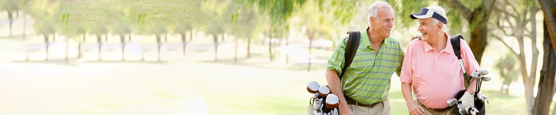Zwei ältere Herren in sommerlicher Kleidung stehen entspannt auf einem sonnigen Golfplatz und unterhalten sich herzlich miteinander. Der eine trägt ein grün-weiß gestreiftes Poloshirt, der andere ein rosa Poloshirt und eine Baseballkappe. Beide haben ihre Golfbags dabei und lächeln sich strahlend an, während sie offensichtlich einen vergnüglichen Moment beim gemeinsamen Golfspielen erleben. Die warme, helle Atmosphäre und ihre gelöste Körperhaltung vermitteln pure Lebensfreude und Unbeschwertheit. Das Bild strahlt aus, dass auch im fortgeschrittenen Alter aktive Freizeitgestaltung und gesellige Momente mit Freunden ohne Sorgen und Einschränkungen möglich sind - ein Gefühl von Freiheit und Selbstvertrauen, das durch die richtige Unterstützung bei Kontinenzproblemen erreicht werden kann.