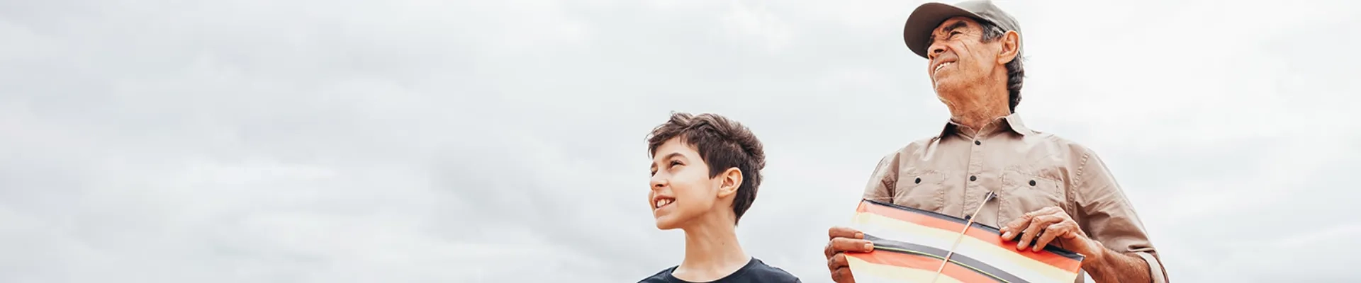 Ein älterer Mann mit grauer Schirmmütze und beigefarbenem Hemd steht entspannt neben einem jungen Jungen mit dunklen, lockigen Haaren und dunklem T-Shirt. Beide lächeln strahlend und blicken hoffnungsvoll in die Ferne, während der Mann einen bunten Drachen in den Händen hält. Der weite, helle Himmel im Hintergrund verstärkt das Gefühl von Freiheit und Unbeschwertheit. Die Szene vermittelt pure Lebensfreude und zeigt, wie Großvater und Enkel einen wunderschönen Moment miteinander teilen – ein Bild für die schönen Dinge des Lebens, die man ohne Sorgen und mit vollem Vertrauen genießen kann.