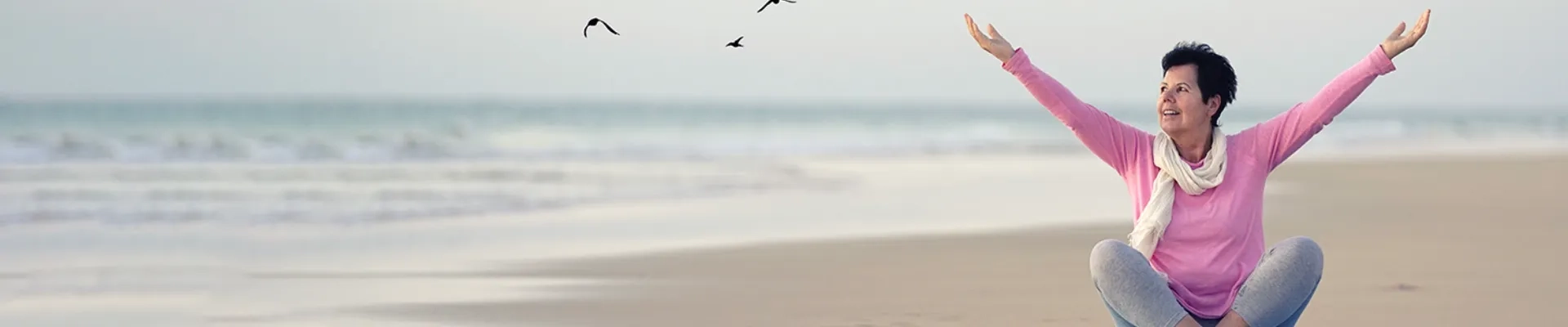 Eine Frau mittleren Alters sitzt entspannt im Schneidersitz am Strand und streckt beide Arme freudig in die Luft. Sie trägt einen rosafarbenen Pullover und einen hellen Schal, ihr Gesicht strahlt pure Lebensfreude und Zufriedenheit aus. Hinter ihr erstreckt sich ein weiter, ruhiger Strand mit sanften Wellen, über dem Möwen durch den hellen Himmel gleiten. Die gesamte Szenerie vermittelt ein Gefühl von Freiheit, Unbeschwertheit und wiedergewonnener Lebensqualität - eine Frau, die das Leben in vollen Zügen genießt und sich keine Sorgen macht, sondern voller Vertrauen und Gelassenheit den Moment auskosten kann.