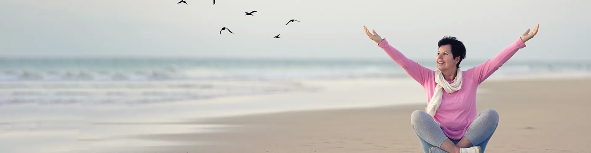Eine Frau mittleren Alters sitzt entspannt im Schneidersitz am Strand und streckt beide Arme freudig in die Luft. Sie trägt einen rosafarbenen Pullover und einen hellen Schal, ihr Gesicht strahlt pure Lebensfreude und Zufriedenheit aus. Hinter ihr erstreckt sich ein weiter, ruhiger Strand mit sanften Wellen, über dem Möwen durch den hellen Himmel gleiten. Die gesamte Szenerie vermittelt ein Gefühl von Freiheit, Unbeschwertheit und wiedergewonnener Lebensqualität - eine Frau, die das Leben in vollen Zügen genießt und sich keine Sorgen macht, sondern voller Vertrauen und Gelassenheit den Moment auskosten kann.