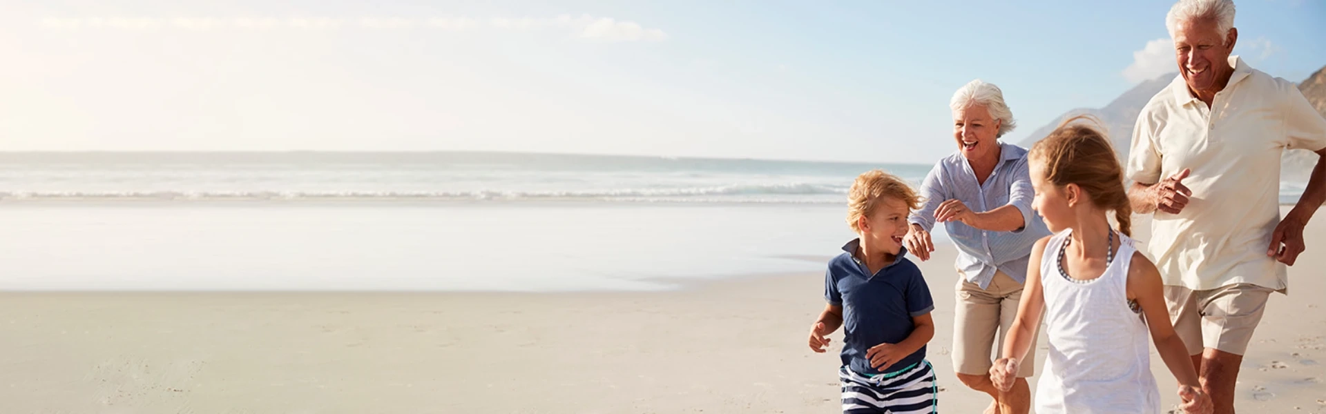 Ein älteres Ehepaar läuft fröhlich und unbeschwert mit ihren beiden Enkelkindern am Strand entlang. Die Großeltern, etwa 65-70 Jahre alt, strahlen vor Freude und Vitalität - die Großmutter trägt eine hellblaue Bluse, der Großvater ein weißes Hemd. Die Kinder, ein Junge in gestreiftem Shirt und ein Mädchen in weißem Top, rennen ausgelassen vor ihnen her. Das warme Sonnenlicht taucht die Szene in goldenes Licht, während sanfte Wellen im Hintergrund an den weiten Sandstrand rollen. Die Familie wirkt völlig entspannt und genießt sichtlich ihre gemeinsame Zeit - ein Bild von Lebensfreude, Aktivität und generationenübergreifender Verbundenheit. Die Szene vermittelt das Gefühl von Sicherheit und Unbeschwertheit, als könnten die Großeltern ohne jede Sorge alle Momente mit ihren Liebsten voll auskosten.