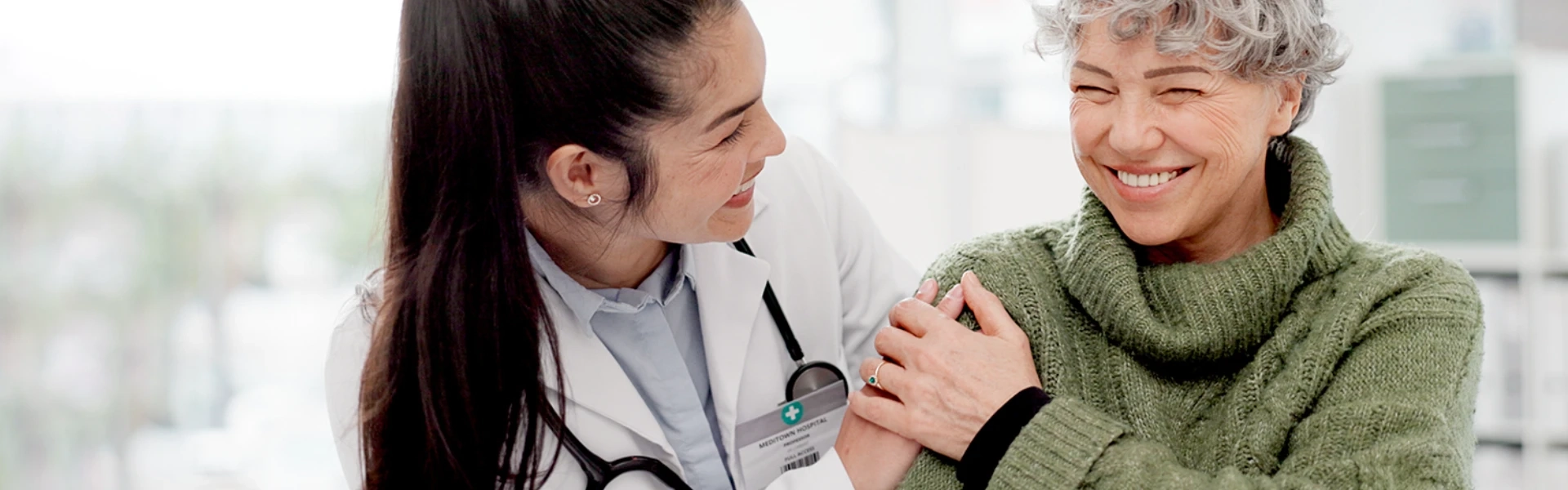 Eine junge Ärztin mit dunklen, zu einem Zopf gebundenen Haaren und weißem Kittel wendet sich warmherzig einer älteren Patientin zu. Die Patientin, eine Frau mit kurzen, silbergrauen Locken, trägt einen gemütlichen olivgrünen Strickpullover und strahlt voller Lebensfreude. Beide Frauen lächeln einander herzlich an - die Ärztin mit fürsorglicher Zuwendung, die Patientin mit einem strahlenden, selbstbewussten Lächeln, das pure Lebensfreude ausstrahlt. Die Patientin hat ihre Hand vertrauensvoll auf die Schulter der Ärztin gelegt, was die innige, vertrauensvolle Beziehung zwischen ihnen unterstreicht. Im Hintergrund ist eine helle, freundliche Praxisumgebung zu erkennen. Das Bild vermittelt Geborgenheit, Vertrauen und vor allem die Botschaft, dass ein erfülltes, sorgenfreies Leben trotz gesundheitlicher Herausforderungen möglich ist - die ältere Dame wirkt selbstsicher, aktiv und voller Optimismus.