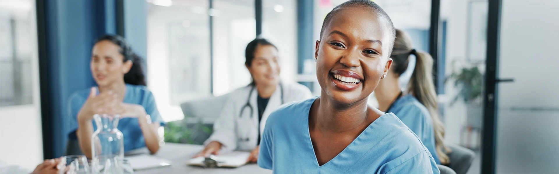 Eine junge, lächelnde Krankenschwester mit kurzen Haaren und hellblauer Arbeitskleidung steht strahlend im Vordergrund eines modernen Schulungsraums. Ihr offenes, herzliches Lachen und ihre entspannte Körperhaltung vermitteln Selbstvertrauen und Professionalität. Im Hintergrund sitzen weitere Kollegen in medizinischer Arbeitskleidung an einem Tisch - eine Frau in Blau und ein Arzt im weißen Kittel - die ebenfalls freundlich und engagiert wirken. Die helle, einladende Atmosphäre des Raums mit großen Fenstern unterstreicht das Gefühl von Kompetenz und Vertrauen. Das Bild strahlt aus: Hier arbeiten qualifizierte Fachkräfte, die durch fundierte Schulungen bestens darauf vorbereitet sind, Menschen mit Kontinenzproblemen einfühlsam und professionell zu betreuen - für ein Leben voller Würde und Unbeschwertheit.