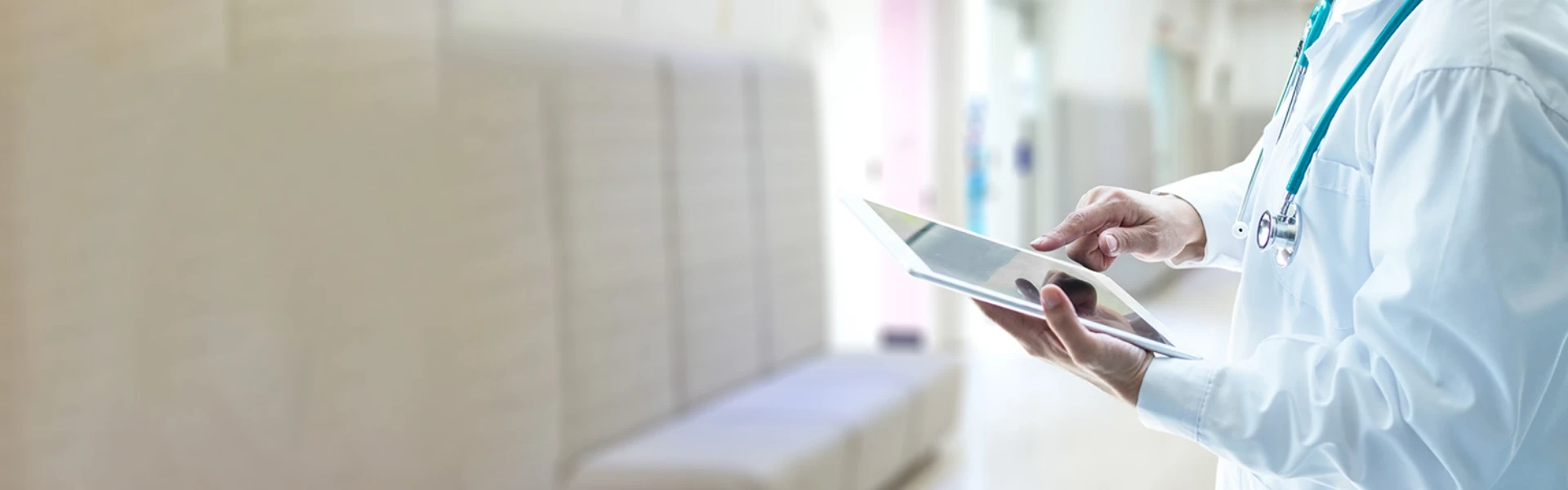 Ein Arzt in weißem Kittel und umhängenden Stetoskop tippt auf einem Tablet herum. Im Hintergrund ist ein Praxis-Wartebereich zu sehen.