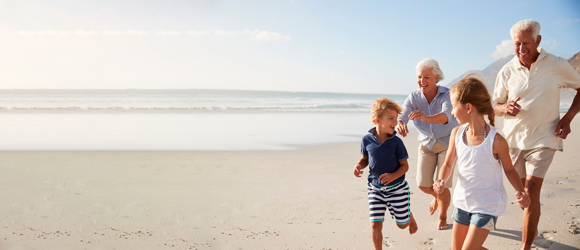 Ein älteres Ehepaar läuft fröhlich und unbeschwert mit ihren beiden Enkelkindern am Strand entlang. Die Großeltern, etwa 65-70 Jahre alt, strahlen vor Freude und Vitalität - die Großmutter trägt eine hellblaue Bluse, der Großvater ein weißes Hemd. Die Kinder, ein Junge in gestreiftem Shirt und ein Mädchen in weißem Top, rennen ausgelassen vor ihnen her. Das warme Sonnenlicht taucht die Szene in goldenes Licht, während sanfte Wellen im Hintergrund an den weiten Sandstrand rollen. Die Familie wirkt völlig entspannt und genießt sichtlich ihre gemeinsame Zeit - ein Bild von Lebensfreude, Aktivität und generationenübergreifender Verbundenheit. Die Szene vermittelt das Gefühl von Sicherheit und Unbeschwertheit, als könnten die Großeltern ohne jede Sorge alle Momente mit ihren Liebsten voll auskosten.