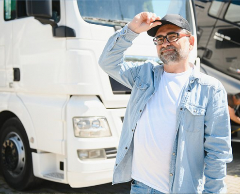Ein selbstbewusster Lkw-Fahrer mittleren Alters mit grauem Vollbart und Brille steht entspannt vor seinem weißen Sattelschlepper. Er trägt eine hellblaue Jeansjacke über einem weißen T-Shirt und eine graue Baseballkappe, die er lässig mit einer Hand zurechtrückt. Sein warmes Lächeln und seine gelöste Körperhaltung strahlen beruflichen Stolz und Zufriedenheit aus. Im Hintergrund steht sein gepflegter Lastwagen, der seine Arbeitswelt und Mobilität repräsentiert. Die Szene vermittelt das Gefühl von Kompetenz und Selbstvertrauen – ein erfahrener Berufskraftfahrer, der seinen anspruchsvollen Job mit Leidenschaft ausübt. Das Bild zeigt, dass Truckerfahrer mit Kontinenzproblemen dank zuverlässiger Hilfsmittel ihren Beruf uneingeschränkt und sorgenfrei ausüben können, ohne sich Gedanken über lange Fahrstrecken oder eingeschränkte Pausenmöglichkeiten machen zu müssen.