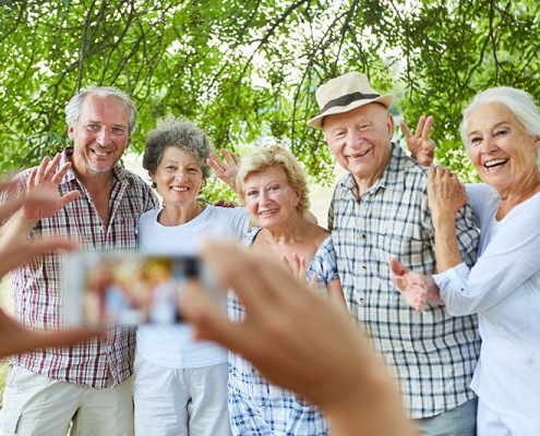 Eine fröhliche Gruppe von fünf älteren Familienmitgliedern und Freunden posiert strahlend für ein gemeinsames Foto unter dem schützenden Blätterdach majestätischer Bäume. Die liebevoll zusammenstehenden Menschen zwischen 60 und 75 Jahren winken herzlich in die Kamera, ihre Gesichter leuchten vor Glück und Verbundenheit. Ein Mann mit Panama-Hut und kariertem Hemd legt seinen Arm um eine Frau mit silbergrauen Locken, während die anderen sich eng aneinander schmiegen - ein Bild von Vertrauen und familiärer Wärme. Ihre sommerliche Kleidung in hellen, freundlichen Farben unterstreicht die unbeschwerte Atmosphäre dieses besonderen Moments. Im Vordergrund sieht man verschwommen die Hände der Person, die das Foto macht, was die intime, persönliche Stimmung dieses Familientreffens verstärkt.