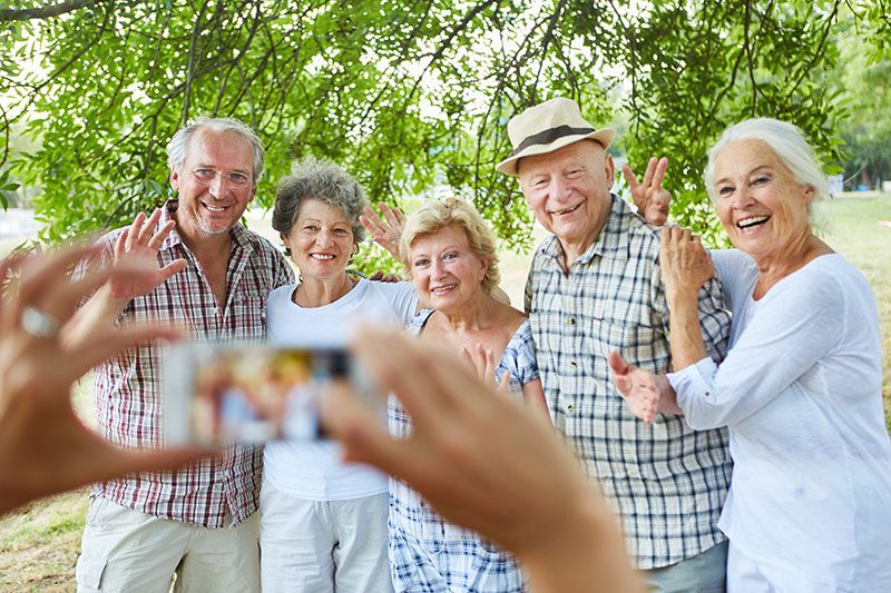 Eine fröhliche Gruppe von fünf älteren Familienmitgliedern und Freunden posiert strahlend für ein gemeinsames Foto unter dem schützenden Blätterdach majestätischer Bäume. Die liebevoll zusammenstehenden Menschen zwischen 60 und 75 Jahren winken herzlich in die Kamera, ihre Gesichter leuchten vor Glück und Verbundenheit. Ein Mann mit Panama-Hut und kariertem Hemd legt seinen Arm um eine Frau mit silbergrauen Locken, während die anderen sich eng aneinander schmiegen - ein Bild von Vertrauen und familiärer Wärme. Ihre sommerliche Kleidung in hellen, freundlichen Farben unterstreicht die unbeschwerte Atmosphäre dieses besonderen Moments. Im Vordergrund sieht man verschwommen die Hände der Person, die das Foto macht, was die intime, persönliche Stimmung dieses Familientreffens verstärkt.