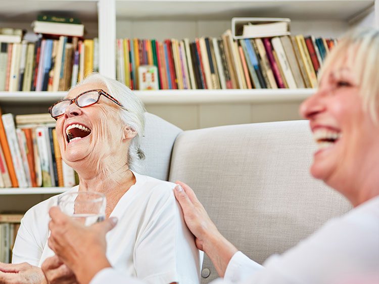 An alte Frau und ihre Tochter sitzen entspannt auf einem hellen Sofa in ihrem gemütlichen Wohnzimmer und lachen herzlich zusammen. Die Frau mit silbergrauen Haaren und Brille hat den Kopf zurückgelehnt und lacht aus vollem Herzen, während ihre Tochter liebevoll ihre Hand auf ihre Schulter gelegt hat und ihr ein Glas Wasser reicht. Im Hintergrund erstreckt sich ein großes Bücherregal voller bunter Bücher. Die Szene strahlt pure Lebensfreude und tiefe Verbundenheit aus.