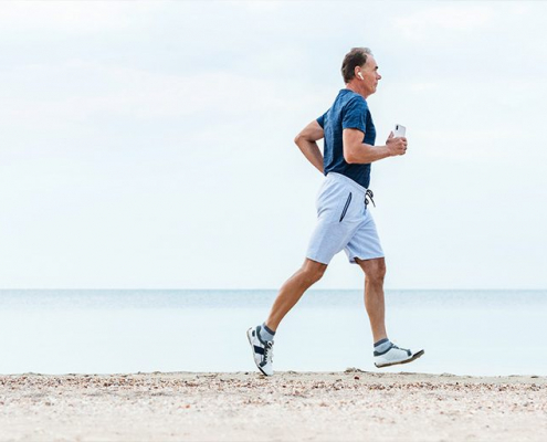 Ein Mann mittleren Alters joggt entspannt und selbstbewusst am Strand entlang. Er trägt ein blaues T-Shirt, helle Shorts und Laufschuhe, während er mit einer Wasserflasche in der Hand seinen Lauf genießt. Im Hintergrund erstreckt sich das ruhige Meer unter einem weiten, hellen Himmel. Seine Körperhaltung strahlt Energie und Lebensfreude aus - er läuft mit aufrechtem Oberkörper und gleichmäßigen Schritten, sein Gesichtsausdruck wirkt gelassen und zufrieden. Das Bild vermittelt ein Gefühl von Unbeschwertheit, Vitalität und dem Vertrauen, aktiv am Leben teilnehmen zu können. Es zeigt, dass körperliche Aktivitäten und Outdoor-Erlebnisse ohne Sorgen oder Einschränkungen möglich sind - ein Symbol für die Freiheit, das Leben in vollen Zügen zu genießen.