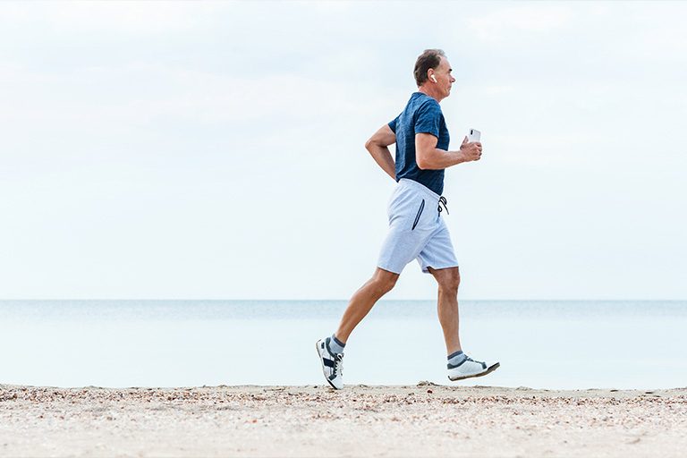 Ein Mann mittleren Alters joggt entspannt und selbstbewusst am Strand entlang. Er trägt ein blaues T-Shirt, helle Shorts und Laufschuhe, während er mit einer Wasserflasche in der Hand seinen Lauf genießt. Im Hintergrund erstreckt sich das ruhige Meer unter einem weiten, hellen Himmel. Seine Körperhaltung strahlt Energie und Lebensfreude aus - er läuft mit aufrechtem Oberkörper und gleichmäßigen Schritten, sein Gesichtsausdruck wirkt gelassen und zufrieden. Das Bild vermittelt ein Gefühl von Unbeschwertheit, Vitalität und dem Vertrauen, aktiv am Leben teilnehmen zu können. Es zeigt, dass körperliche Aktivitäten und Outdoor-Erlebnisse ohne Sorgen oder Einschränkungen möglich sind - ein Symbol für die Freiheit, das Leben in vollen Zügen zu genießen.